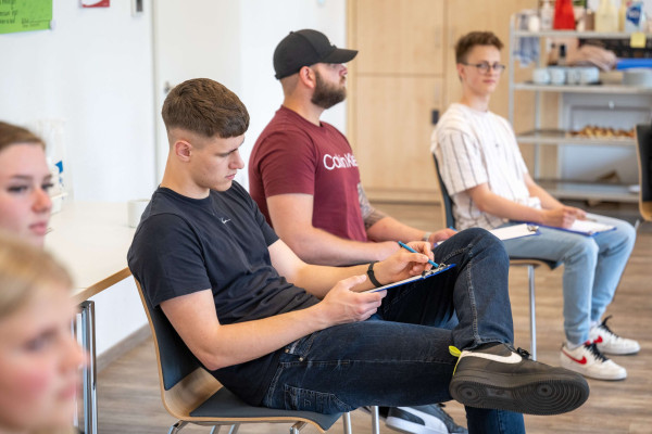 Drei junge Männer sitzen in einer Seminarrunde, einer schreibt auf ein Klemmbrett