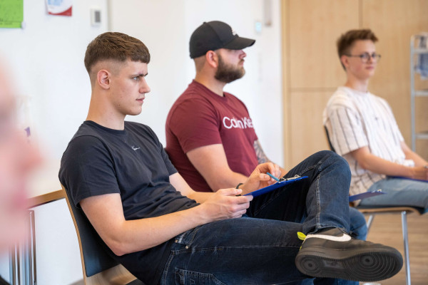 Drei junge Männer sitzen in einer Seminarrunde, einer macht sich Notizen