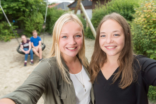 Zwei junge Frauen machen ein Selfie auf einem Spielplatz, im Hintergrund sitzen Kinder in einer Schaukel