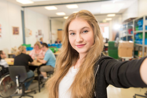 Junge Frau macht ein Selfie in einer Einrichtung, im Hintergrund sitzen Menschen an Tischen
